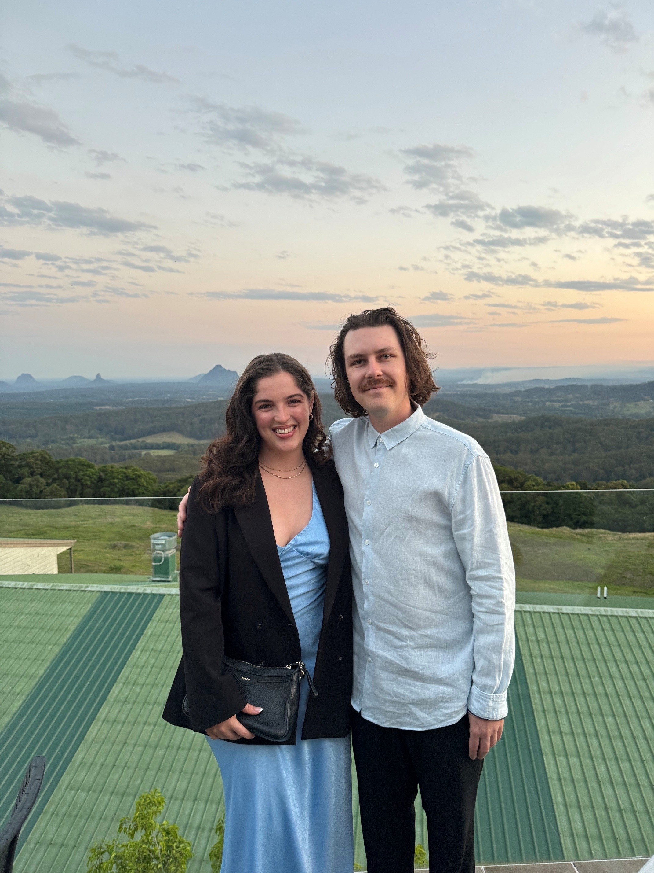 A woman in a blue dress, a black jacket, a man in a pale blue button-down shirt, against the backdrop of mountains at sunset.