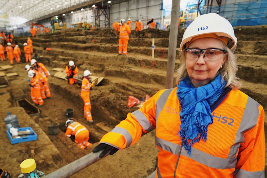 A blonde woman in safety glasses, hard hat and high-vis clothes stands at an excavation site within a building.