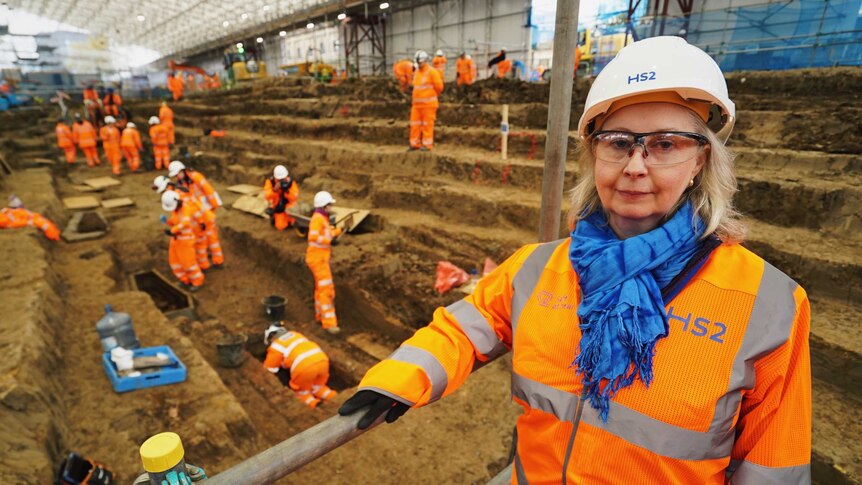 A blonde woman in safety glasses, hard hat and high-vis clothes stands at an excavation site within a building.