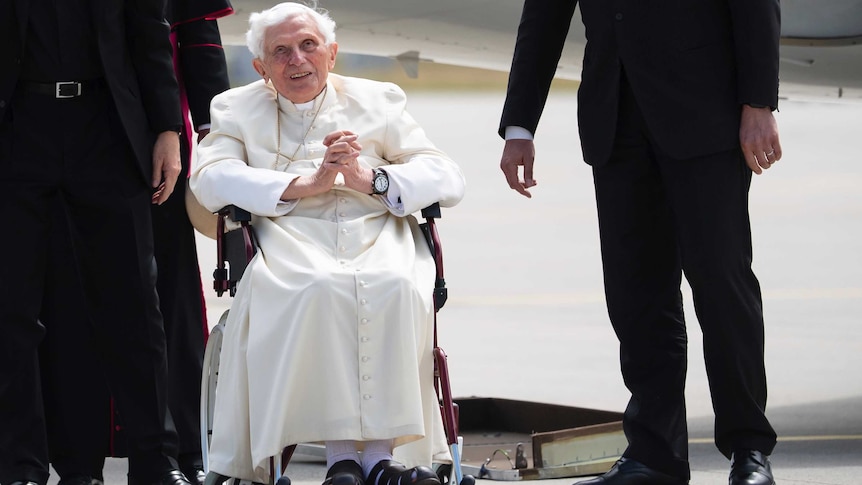 An elderly man wearing white vestments sits in a wheelchair with several other mean wearing black standing around him.