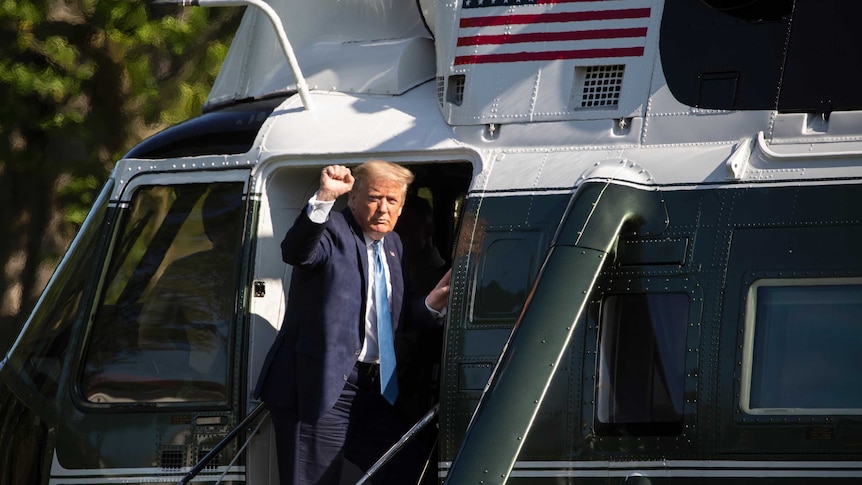 A man in a suit raises his fist as he looks at the camera while leaning out from the door of a helicopter