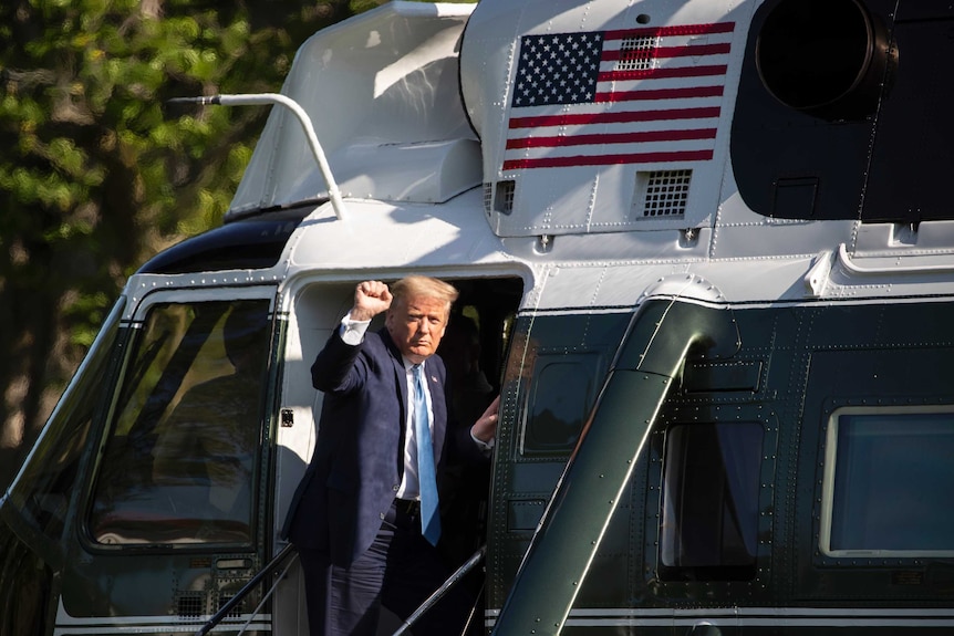 A man in a suit raises his fist as he looks at the camera while leaning out from the door of a helicopter