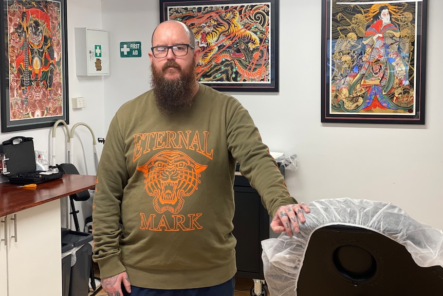 Un hombre barbudo se para en un salón de tatuajes lleno de pinturas coloridas. 