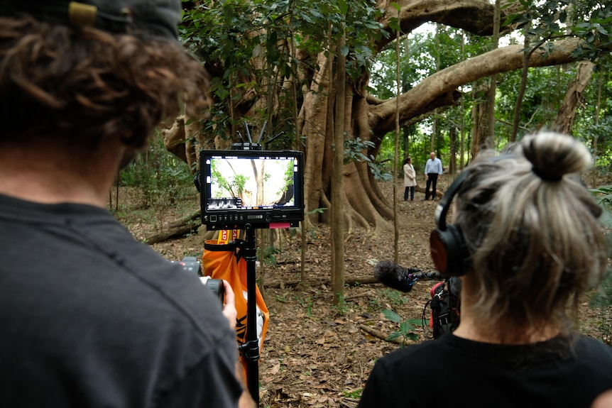Camera crew filming a man and a woman standing in a forest in the distance.