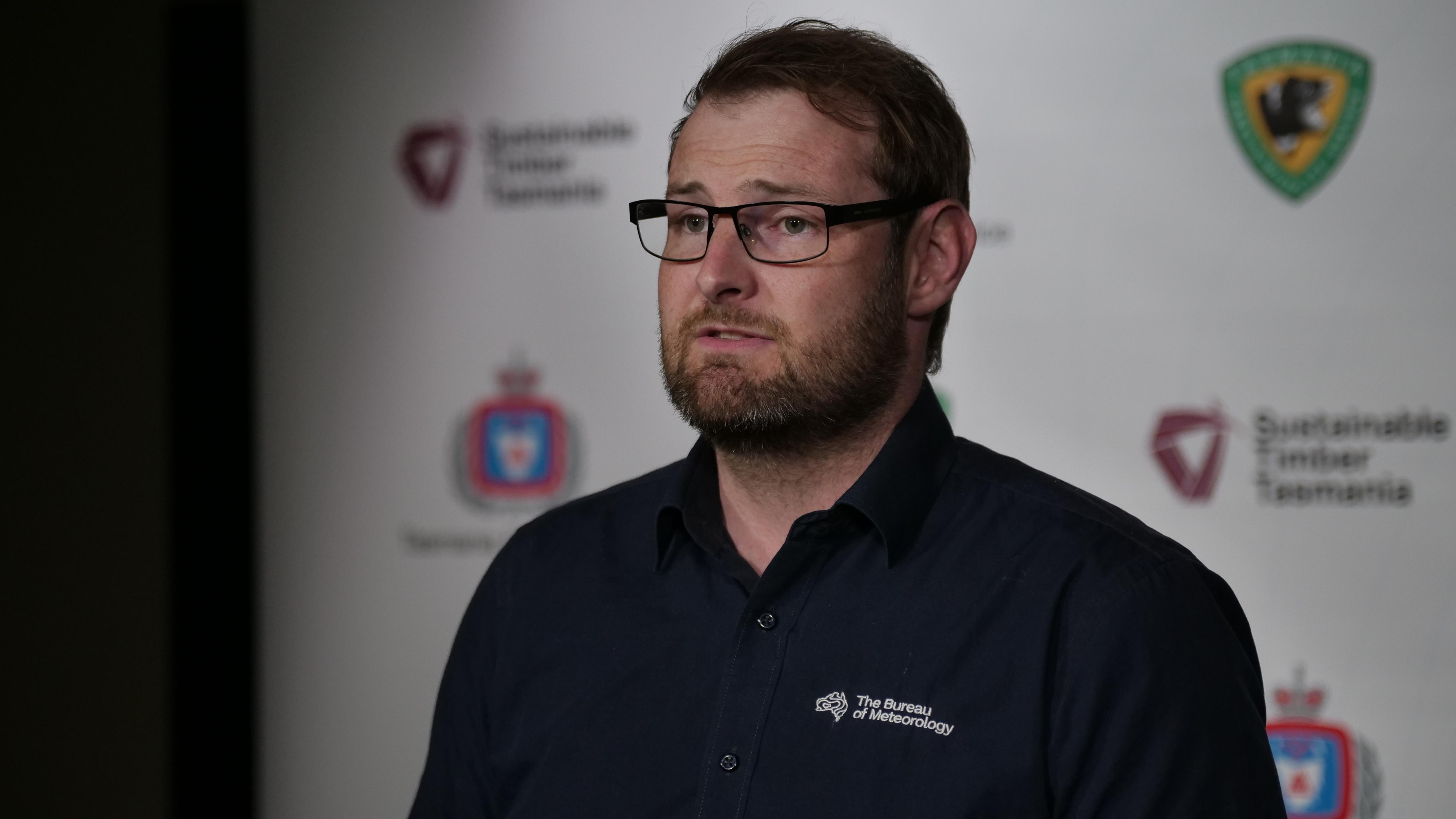 A man in a black polo shirt and black-framed glasses speaks to media. 