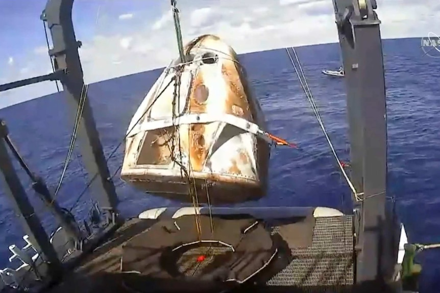 A white triangular space capsule is retrieved from the sea with a custom winch attached to the site of a ship.