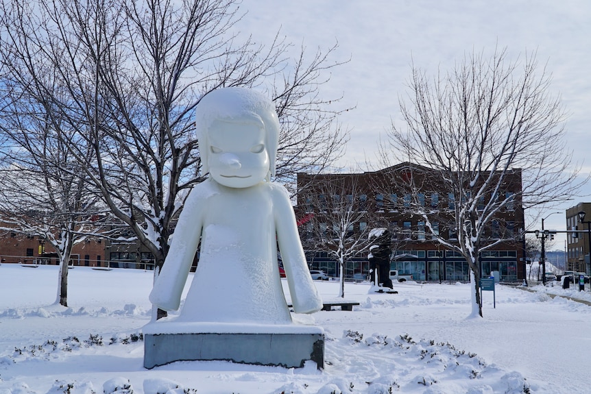 A white sculpture is surrounded by snow. Buildings and skeletal trees are in the background.