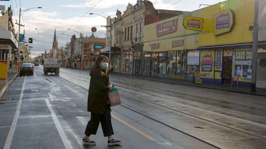A woman wearing a mask crosses the wet road in front of a kebab shop and chemist as the sun beams.
