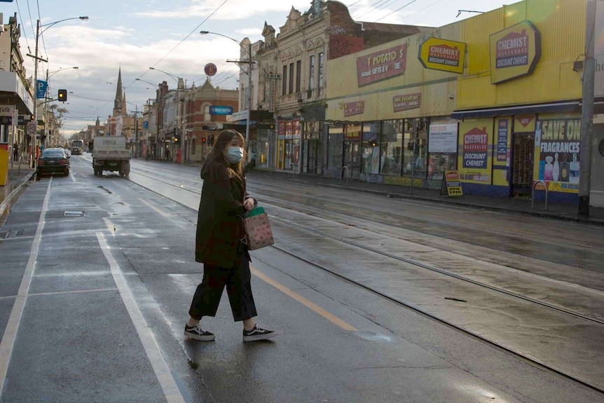 A woman wearing a mask crosses the wet road in front of a kebab shop and chemist as the sun beams.