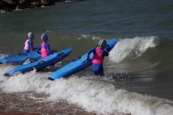 Image of three young life saving club trainees in blue and pink wetsuits with blue bodyboards.