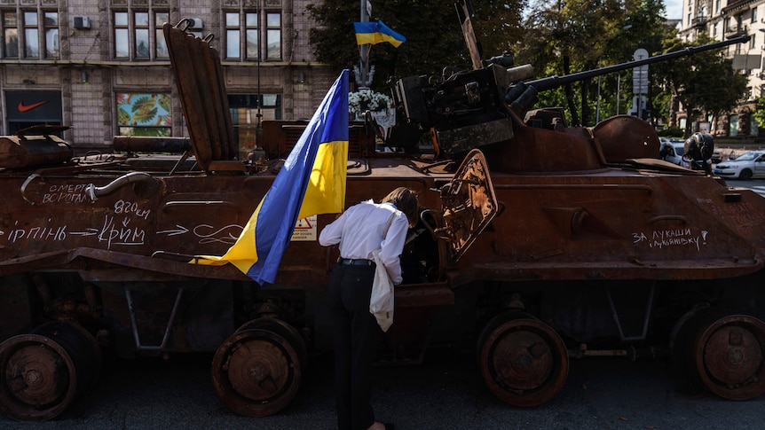 A woman holds the flag of Ukraine while looking into one of the destroyed Russian military vehicles.