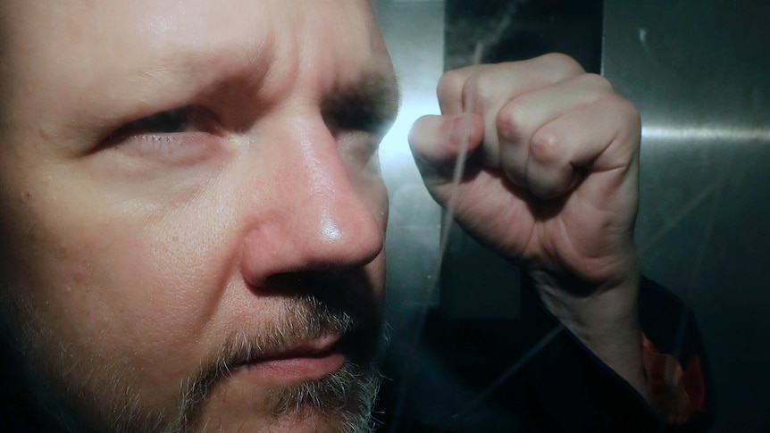 A close up photo of Julian Assange with a beard, while holding his hand in a fist.