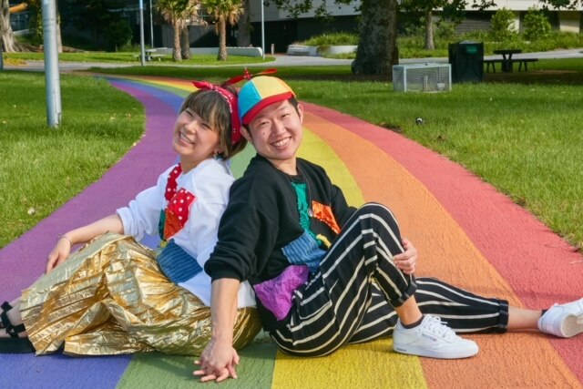 Two young Japanese people are sitting back-to-back on a rainbow path of the ground.