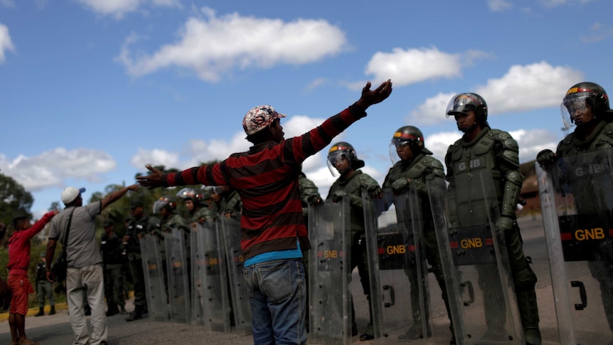 Men gesture at a row of soldiers wearing riot gear who are blocking the Venezuelan border.
