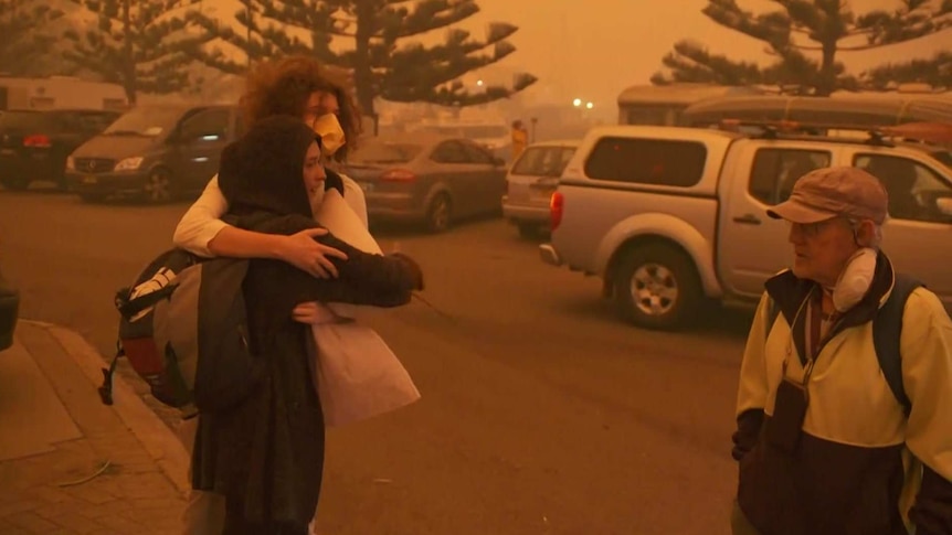 A couple hug as a man passes them in a car park. The sky is orange from the dust in the air.