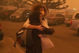 A couple hug as a man passes them in a car park. The sky is orange from the dust in the air.