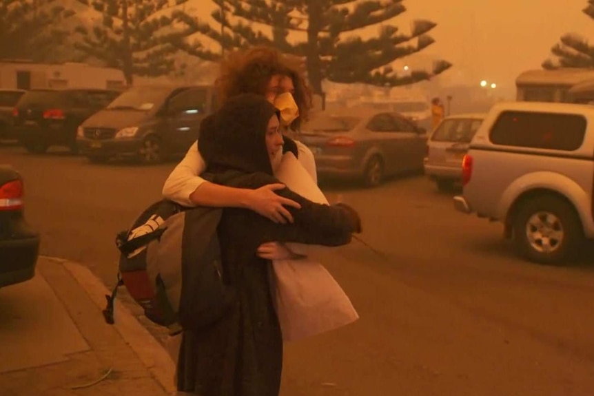 A couple hug as a man passes them in a car park. The sky is orange from the dust in the air.