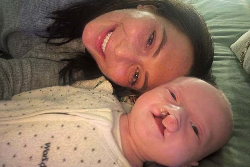 Lana is lying next to her baby. Lana has dark hair and is smiling. The baby has a bilateral cleft palette and is also smiling