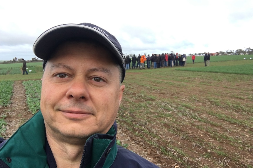 Selfie of a man standing in a paddock, wearing a green & blue jacket, navy cap and a large group of people gathered in distance