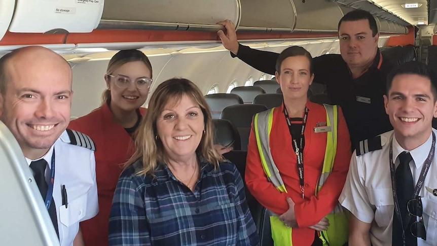 woman in checked shirt surrounded by flight staff on empty aircraft