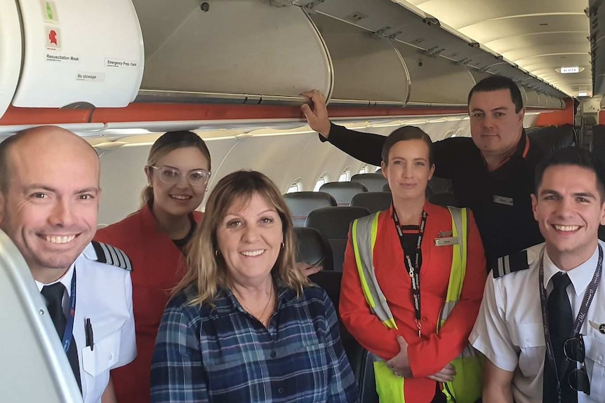 woman in checked shirt surrounded by flight staff on empty aircraft