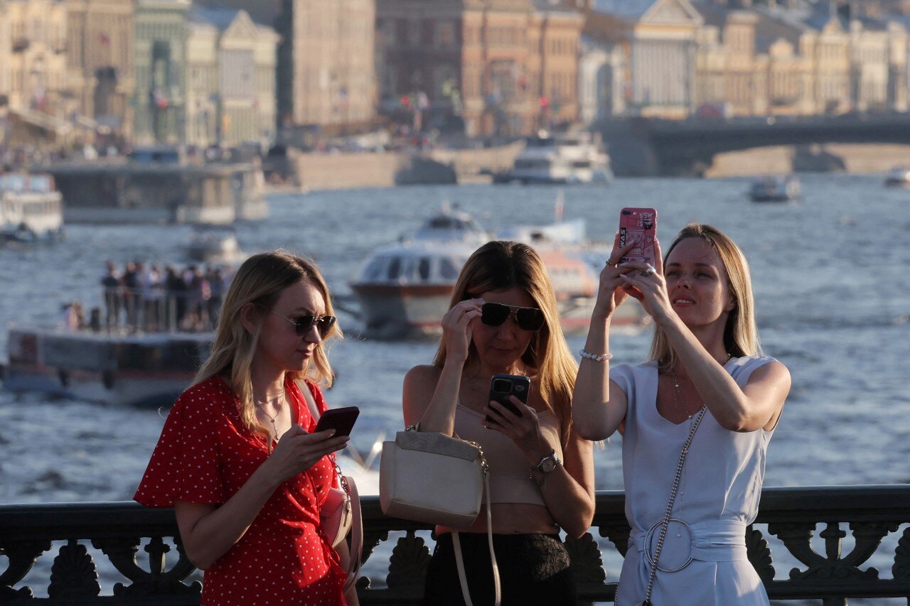 Three women looking at their phones, with water and a city skyline visible behind them.