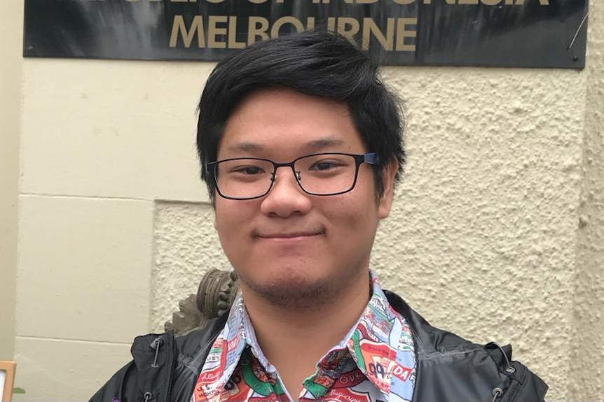 A young man wearing glasses dresses in Indonesian batik standing in front of Melbourne signage
