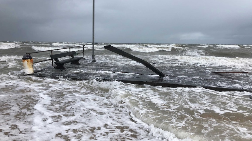 Rough seas wash over wood parts of the Frankston Pier sitting on the sand.