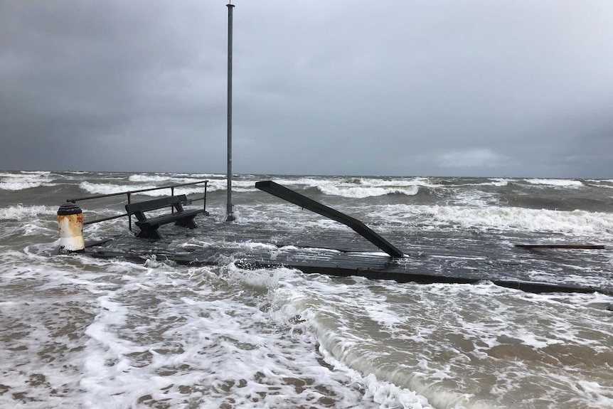 Rough seas wash over wood parts of the Frankston Pier sitting on the sand.