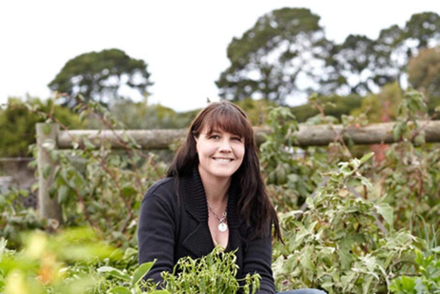 A woman with dark hair crouches in a large vegetable garden, smiling at the camera.
