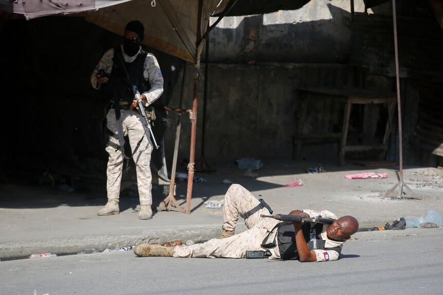 A police man aims a rifle at gang members during a clash. He's laying on his side on the ground.