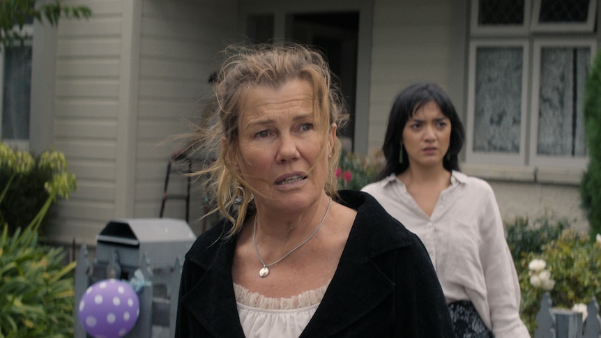 A woman in a black blazer stands in front of a lawn looking concerned, another younger woman behind her also with furrowed brow.