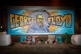 A blue and yellow mural on a wall for George Floyd with bunches of flowers and posters left underneath. 