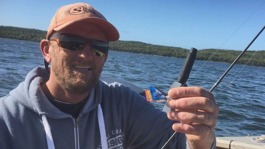 A man wearing a hat and sunglasses sits on a boat holding a fishing rod with a Mentos wrapper tied on as bait.