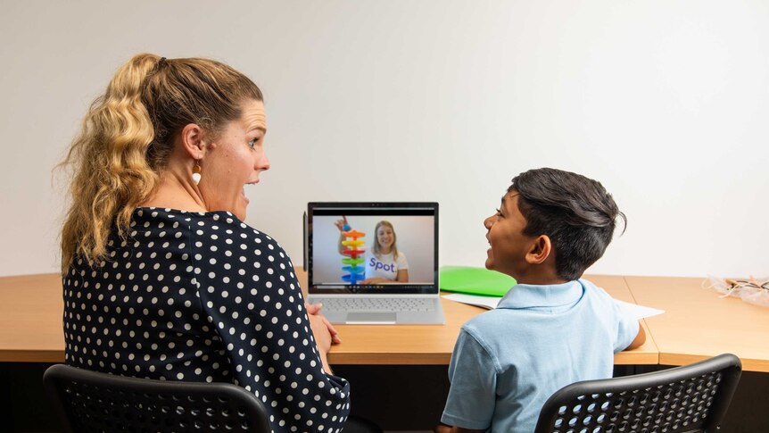 An adult and young child sit in front of a laptop, taking part in an online speech pathology session.