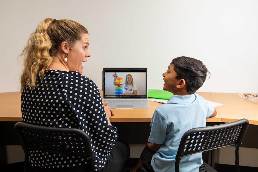 An adult and young child sit in front of a laptop, taking part in an online speech pathology session.