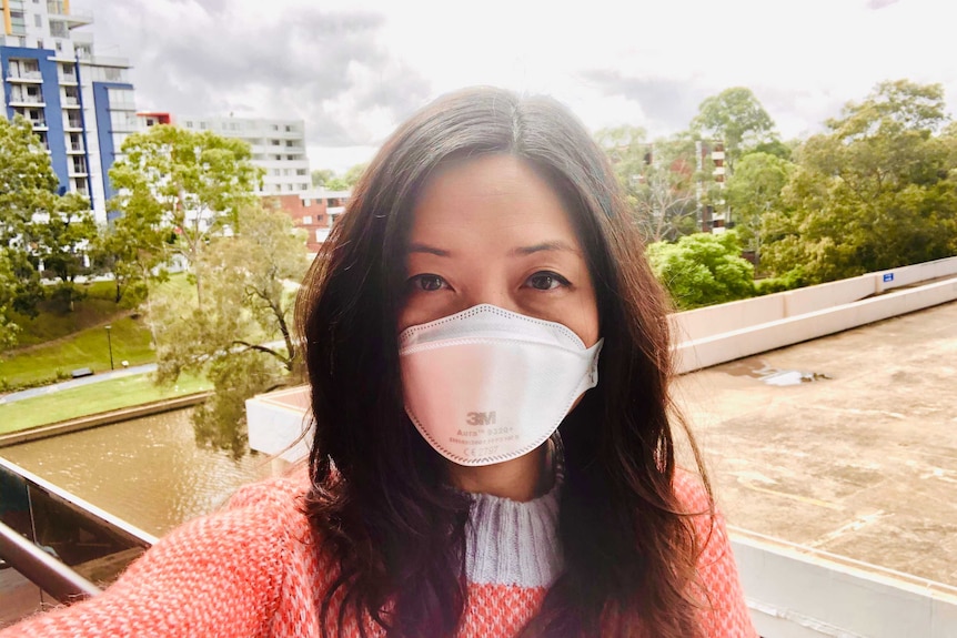 An Asian woman wearing a mask takes a selfie from a balcony with trees and buildings behind her.