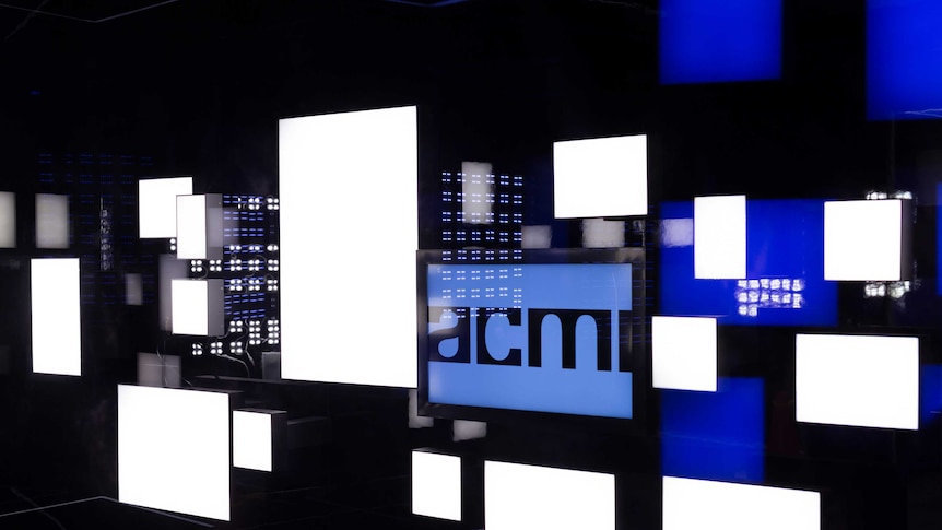 A glass wall with a series of white and blue lit panels. The word ACMI is on a blue frame on the wall.