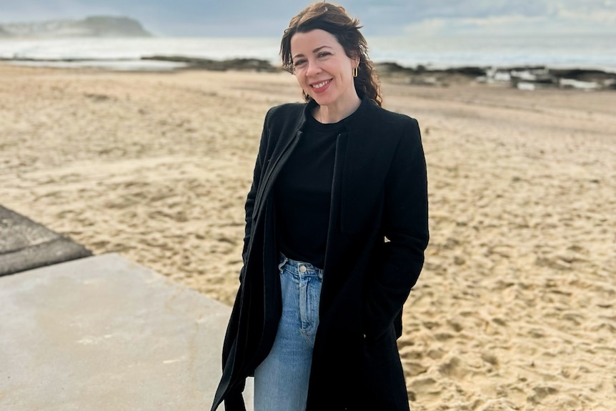 Full body shot of a woman standing by the sand at a beach, wearing a winter coat, with hands in her pockets, smiling.