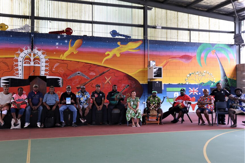 14 community members and elders, most of whom are seated  a brightly coloured wall in an assembly hall.