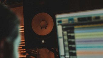 A mixing desk and speaker are in focus, while a man looks at a computer screen showing a music producing program