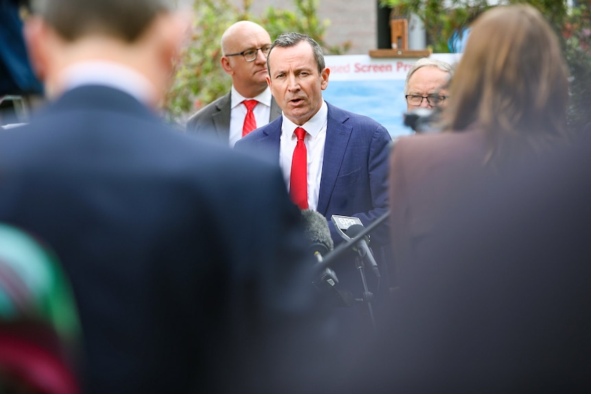 A wide front-on view of Mark McGowan speaking at a media conference outdoors, with people in the foreground.
