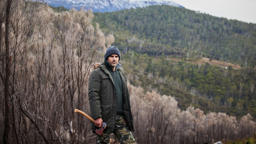 Mike Atkinson wearing camouflage outfit holding an axe in the bush. 