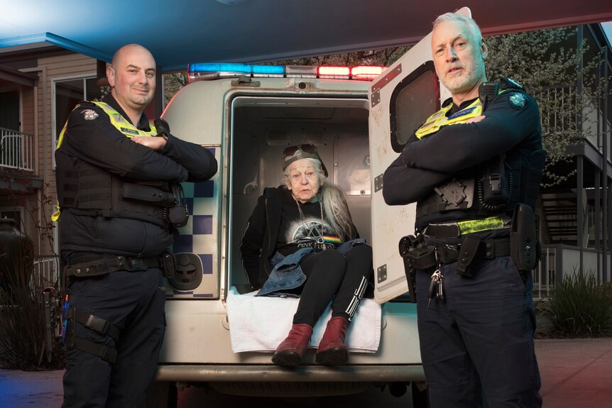 Two police officer stand with arms crossed with an elderly woman in the back of a police van. 