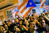 A crowd of people gather at night, waving Puerto Rico flags and chanting.
