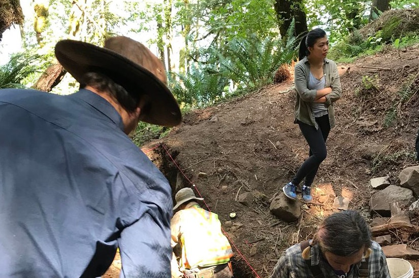 Archaeology students dig in a trench next to lush forest.
