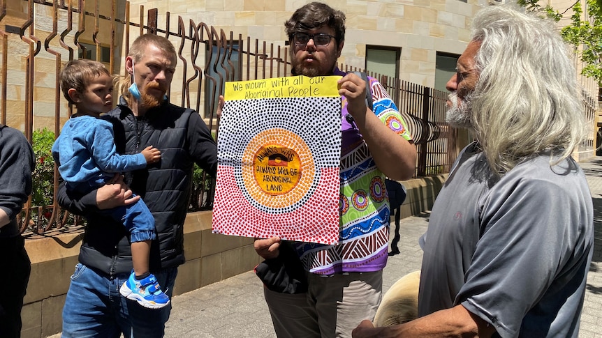 Three men standing outside a court building, one holding a sign, one holding a young child