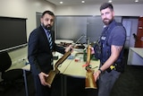Two police officers standing in an office, facing each other while holding old style shot guns, while looking into the camera.