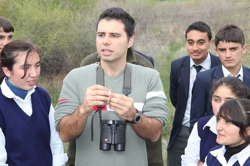 A man holds a small bird in a grassy area as students look on. 