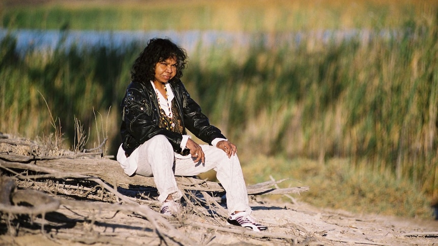 A woman sits on the riverbank looking at the camera. She is wearing a black jacket, a white shirt and white pants.
